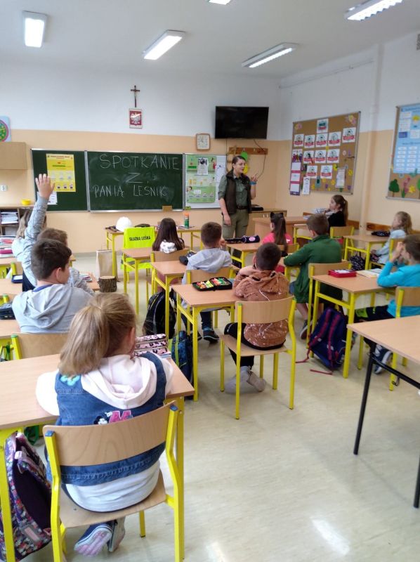 Zdjęcie przedstawia osobę dorosłą oraz dzieci w pomieszczeniu szkolnym.